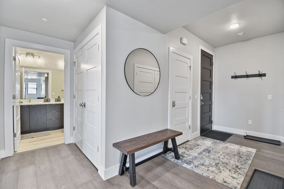 Stylish entryway from the front door into a functional mudroom featuring a wooden bench, wall-mounted coat hooks, and a round mirror. Clean lines, modern doors, and a neutral color palette create a welcoming and practical space for guests.