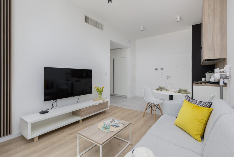 A modern living room with a sleek grey sofa, a coffee table, and a large flat-screen TV. The room is bright, with natural light coming in through floor-to-ceiling windows.