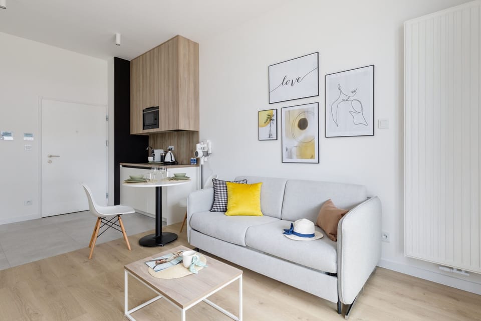 A modern living room with a sleek grey sofa, a coffee table, and a large flat-screen TV. The room is bright, with natural light coming in through floor-to-ceiling windows.