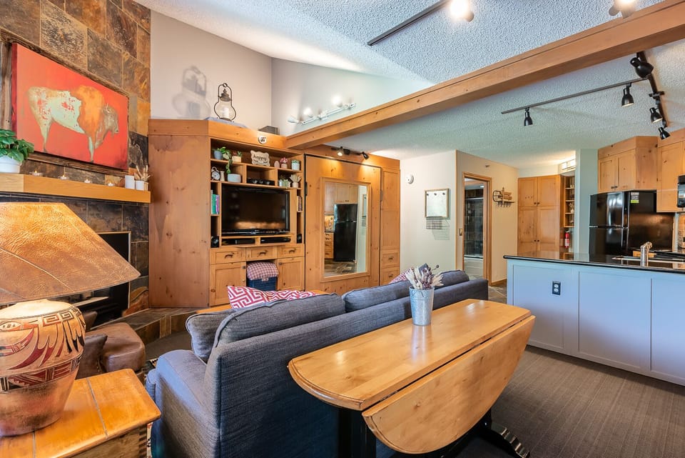 A cozy living room with wooden furniture, a fireplace, a wall-mounted TV, a painting of a cow, and track lighting. An adjoining kitchen features black appliances and an island with a countertop.