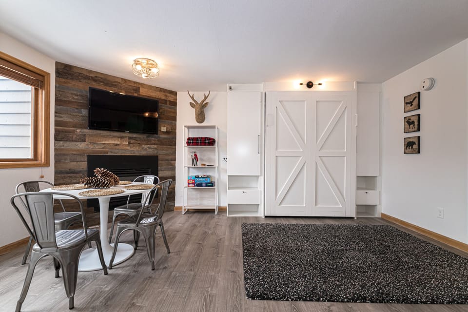 A modern living space with a table and chairs, a mounted TV, a bookshelf, a deer head decoration, a barn style door, and a carpeted area on wooden flooring.