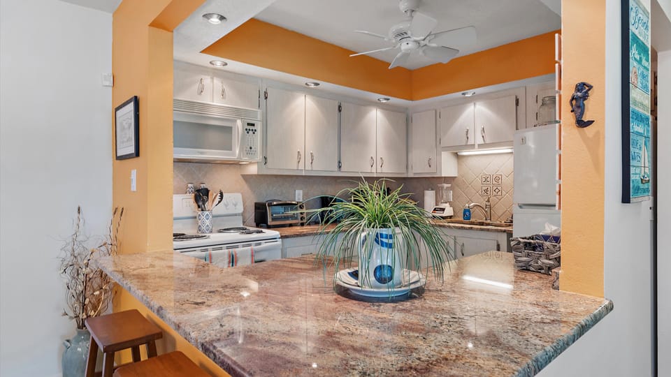 A kitchen with a granite countertop, bar stools, white cabinets, and appliances. A plant in a blue vase sits on the counter, while the walls are painted white and orange. A ceiling fan is also visible.