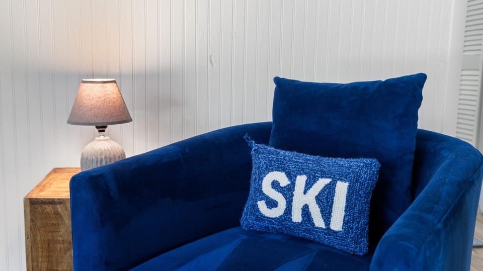 A blue velvet chair with a blue cushion featuring the word "SKI" is placed next to a wooden side table holding a lit lamp. The background consists of white vertical paneling.
