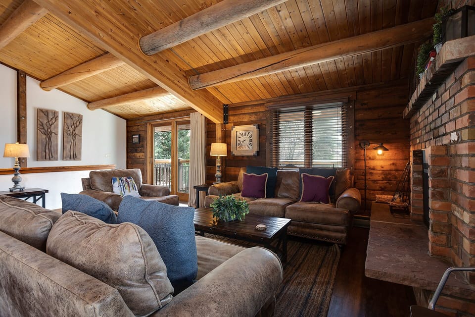 Sink into plush sofas beneath vaulted wood ceilings while natural light pours through your balcony doors. The rustic brick fireplace and warm timber beams create a cozy mountain retreat atmosphere perfect for unwinding.