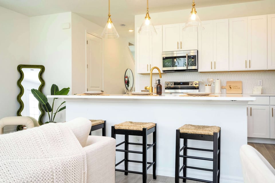 Whip up delicious meals in this bright modern farmhouse kitchen with white shaker cabinets, gold hardware, hex-tile backsplash and three woven-seat bar stools-perfect for casual breakfasts or sunset cocktails!