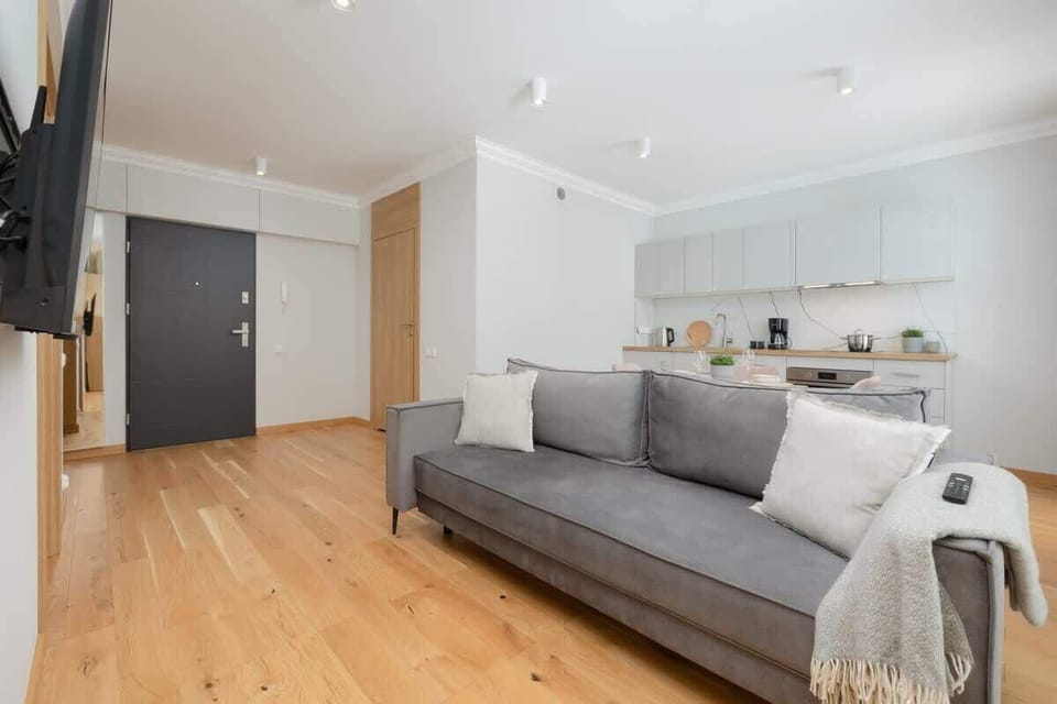 A spacious and bright living room with a gray sofa, light curtains, and wooden flooring, connected to the dining area and kitchen.