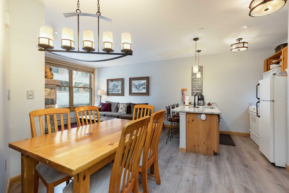 A cozy, modern kitchen and dining area with a wooden dining table, bench, pendant and ceiling lights, and a kitchen island. Decor includes framed art on the walls and a stone accent around the window.