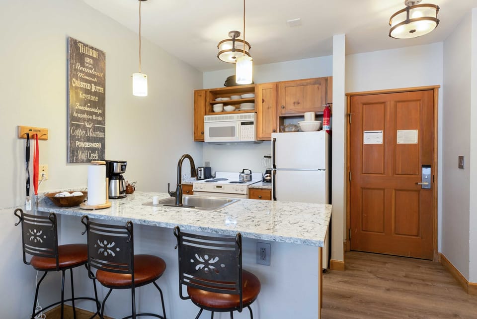 A compact kitchen with a granite counter, three chairs, wooden cabinets, white appliances, and pendant lighting. A sign with inspirational words is on the wall, and keys hang near the door.