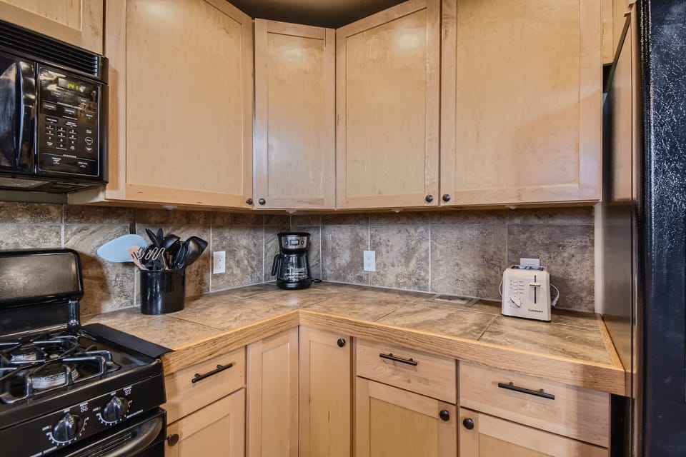 A kitchen with wooden cabinets, a stovetop with black appliances, a coffee maker, a toaster, and a utensil holder on a tiled countertop with a tiled backsplash.
