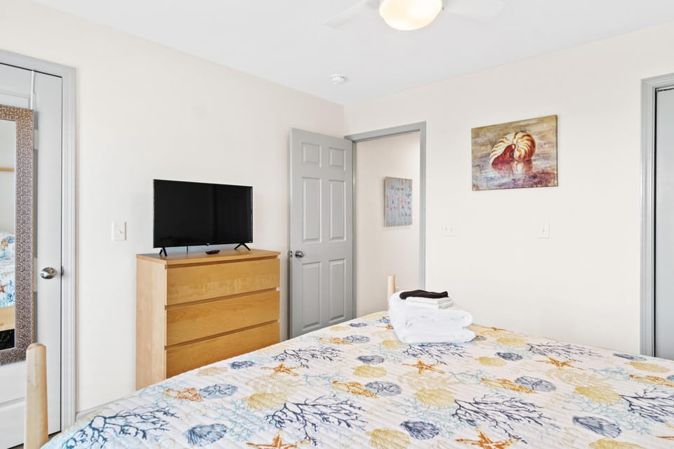 Your comfortable bedroom features a flat-screen TV and dresser, with soft linens and coastal artwork creating a peaceful retreat.