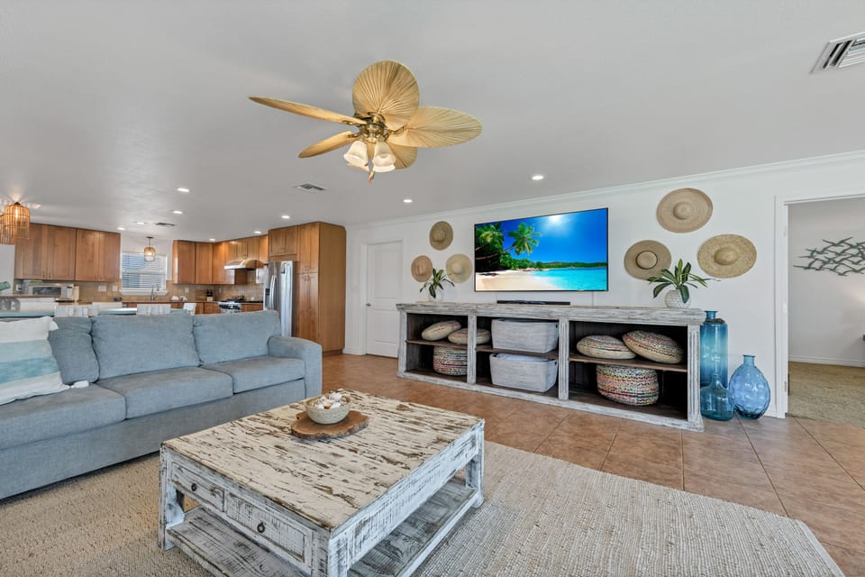 Relax in your open-concept living space with coastal decor, ceiling fan, and tropical TV backdrop creating the perfect beach house vibe.
