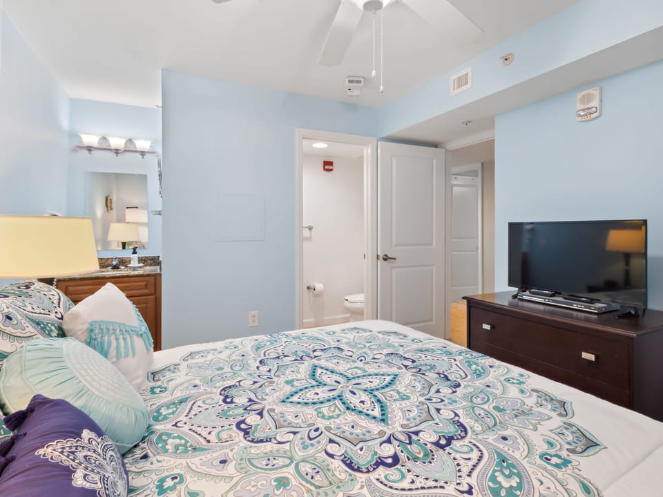 Drift off in your serene bedroom featuring soft blue walls, luxurious bedding with intricate patterns, and modern amenities including flat-screen TV.