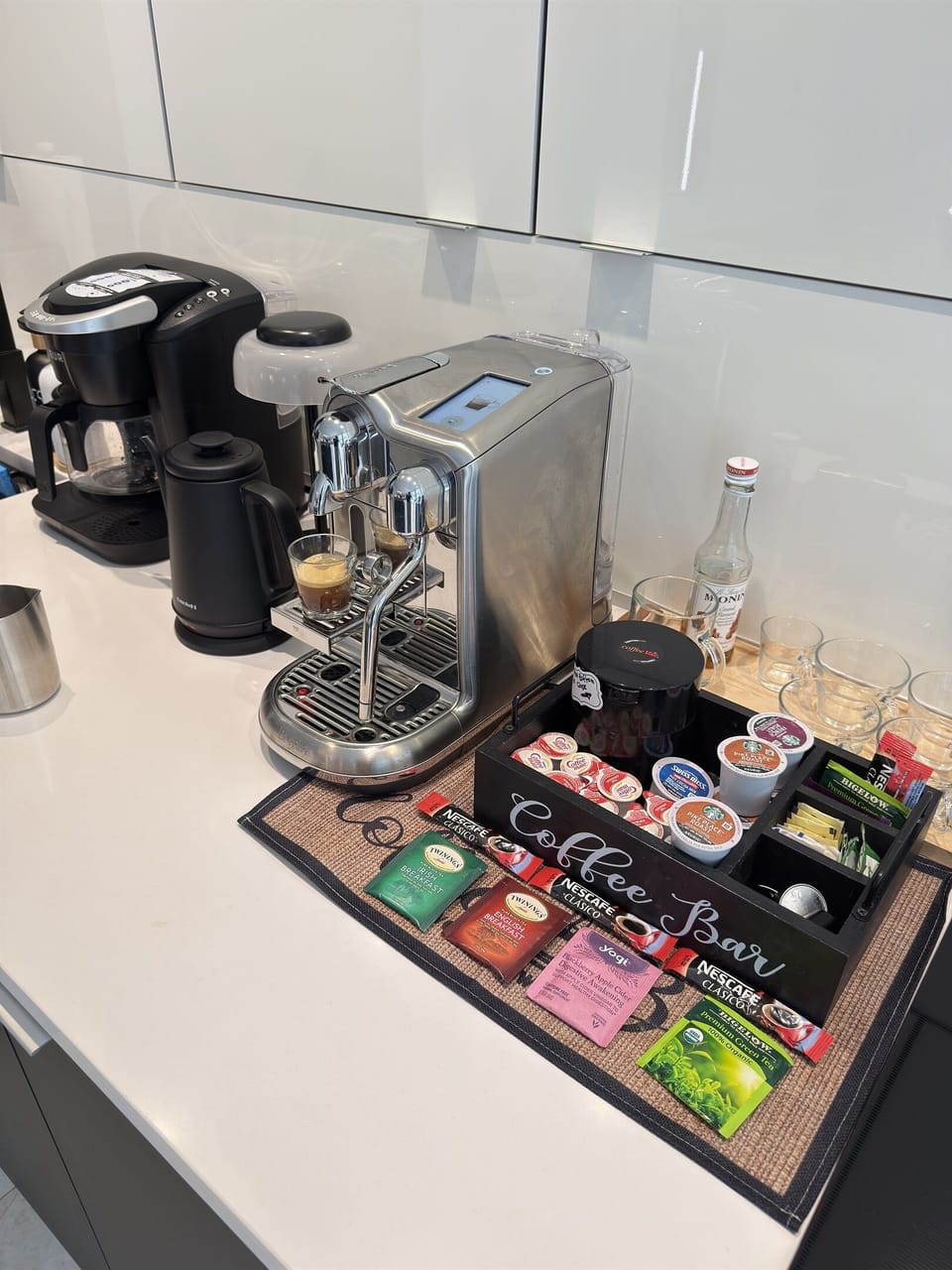 Stylish coffee bar setup featuring a sleek espresso machine, a drip coffee maker, and an organized selection of coffee pods, tea bags, —perfect for any caffeine lover!