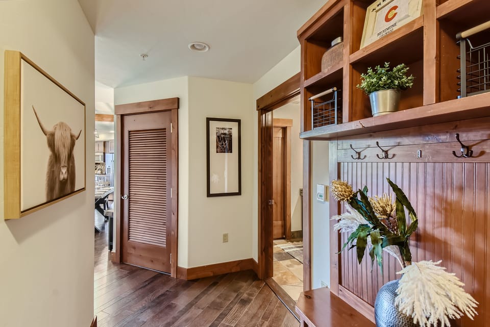 A hallway features wooden accents, a painting of a cow, potted plant, coat hooks, and storage shelves. A louvered door opens to the left, and a glimpse of another room is visible through an open doorway.