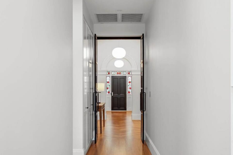 This beautifully composed hallway exudes a sense of understated sophistication. Clean white walls and warm wooden flooring guide the eye toward a stunning black front door, framed by colorful stained glass panels featuring floral motifs. Modern black-fram