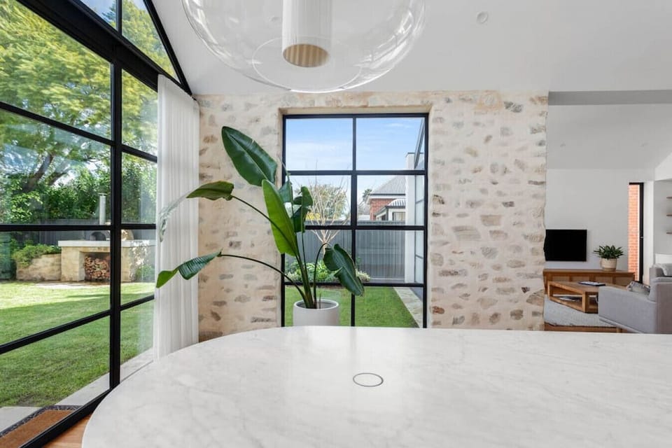 This airy and light-filled interior blends modern elegance with rustic charm. A round marble table anchors the foreground, while large black-framed windows invite abundant natural light and lush garden views into the room. The exposed stone wall adds text