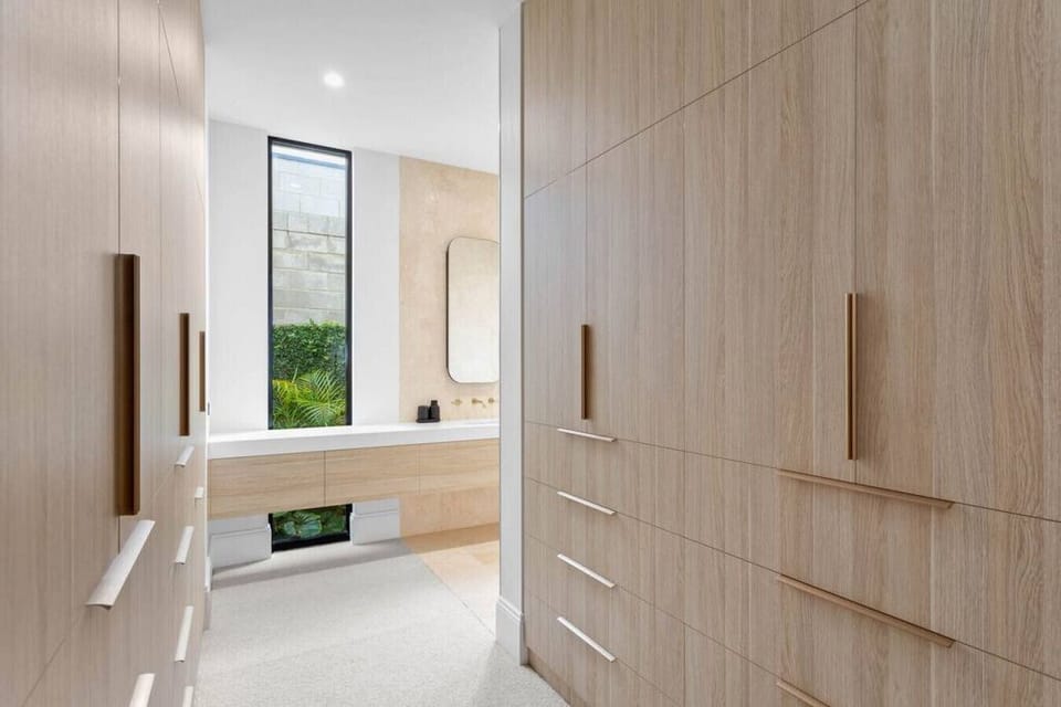 This contemporary walk-in wardrobe exudes elegance and functionality. Flanked by full-height built-in cabinets on both sides, the light wood finishes and gold-toned vertical handles add a refined warmth to the space. The flooring transitions from soft, ne