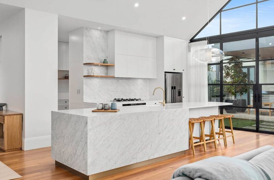 This stunning kitchen features expansive marble benchtops, minimalist white cabinetry, and brushed brass tapware, complemented by natural wood accents. Floor-to-ceiling windows flood the space with light, creating an airy and sophisticated heart of the ho