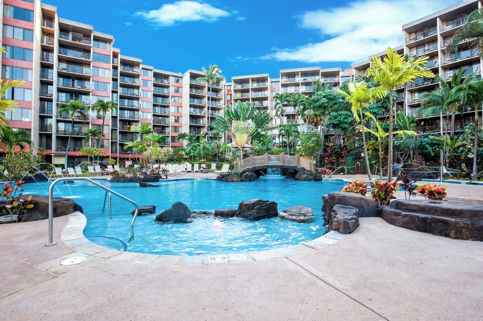 A tropical resort-style pool complex features lush palm trees, natural rock waterfalls, and colorful blooms, set among modern mid-rise buildings under bright blue skies.