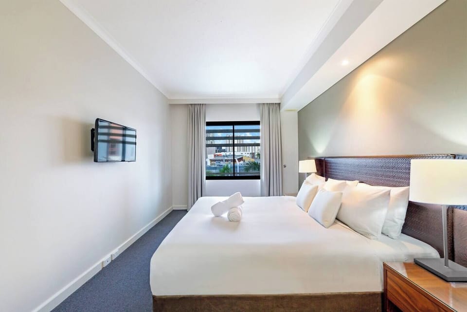 A light-filled bedroom with king bed is complemented by a large window showcasing views of Darwin city and its surroundings.