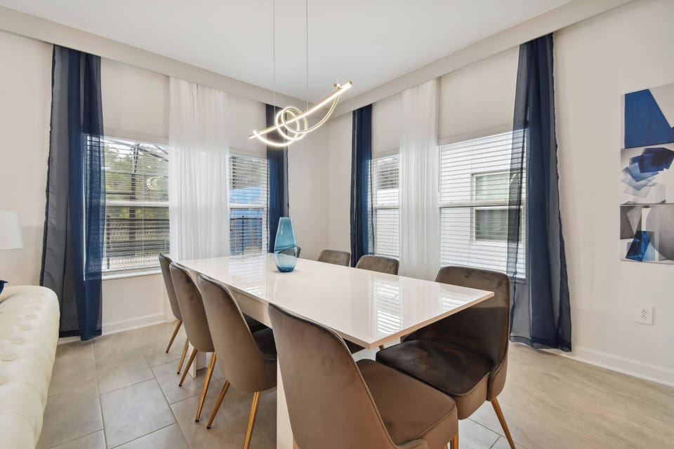 Natural light floods this dining area, highlighting the sleek white table surrounded by stylish, upholstered chairs. The contemporary overhead lighting and blue accents add a touch of modern flair, making every meal feel special.