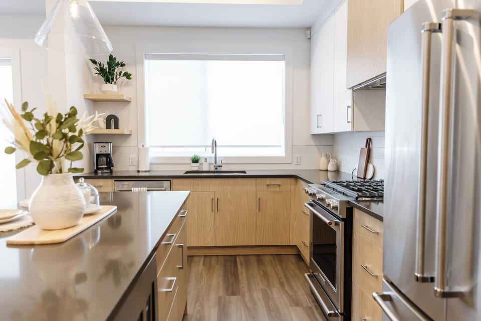Modern kitchen with light wood cabinets, dark countertops, and stainless steel appliances. Enjoy natural light from a large window above the sink, a gas stove, and stylish decor. Perfect for cooking and dining in a bright, welcoming space.