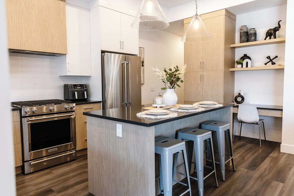 Stylish open-concept kitchen with light wood cabinets, dark granite countertops, and stainless steel appliances. Enjoy a spacious island with seating, pendant lighting, and a cozy workspace, perfect for cooking and relaxing in a modern setting.