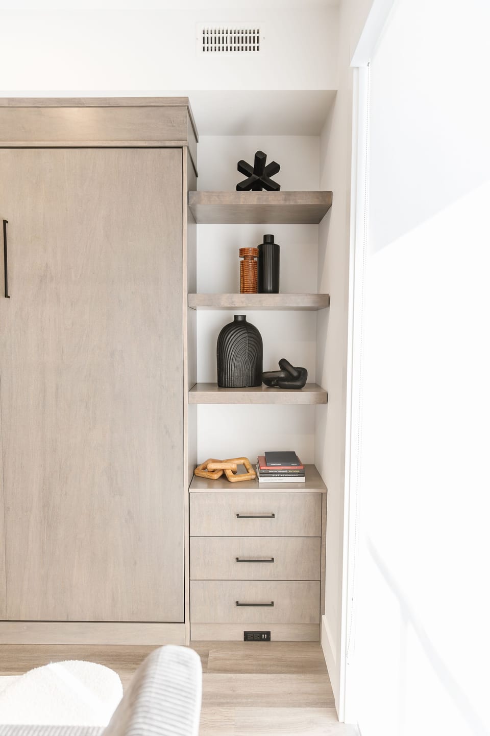 Sleek wall unit with a hidden Murphy bed, open shelves with decorative items, and a small dresser for extra storage. Stylish, modern design with a mix of black and natural wood tones for a cozy, functional space. Perfect for a relaxing stay!