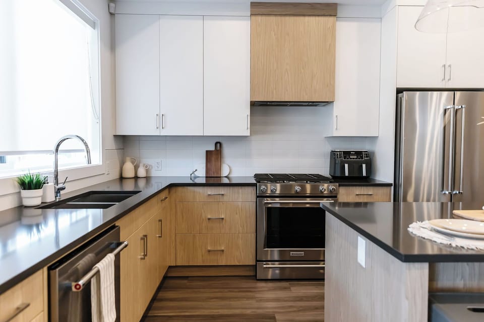 Modern L-shaped kitchen with light wood and white cabinets, dark countertops, and stainless steel appliances. Enjoy a large island, double-basin sink, and natural light from a large window—perfect for cooking and entertaining.