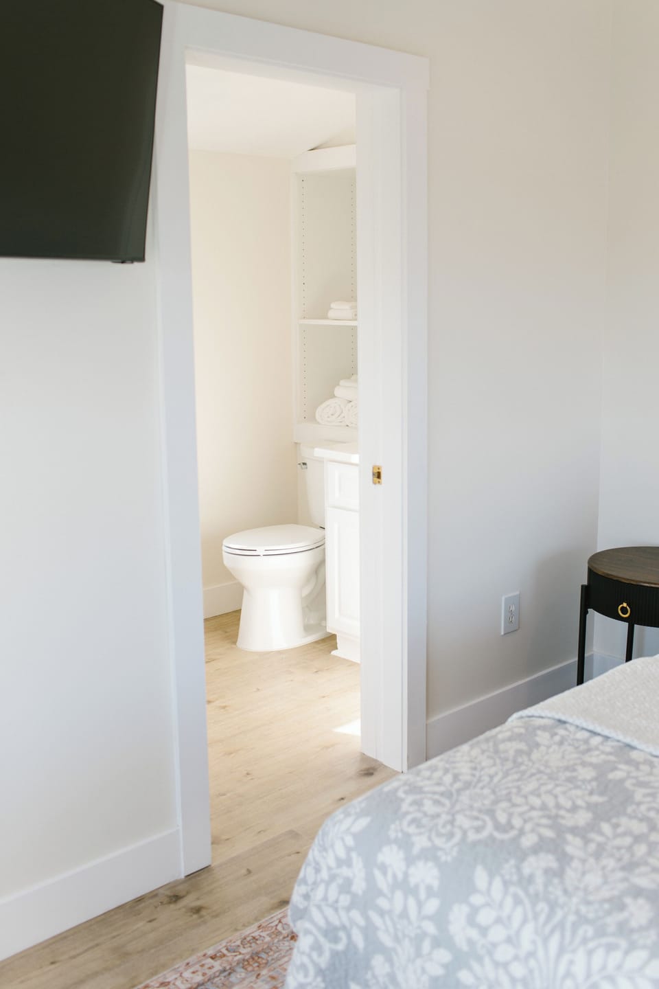 Greenleaf Room 7 features a bright private ensuite with clean white cabinetry, soft wood flooring, and open shelving stocked with plush towels. A seamless blend of comfort and simplicity for your stay in coastal Boothbay Harbor, Maine.