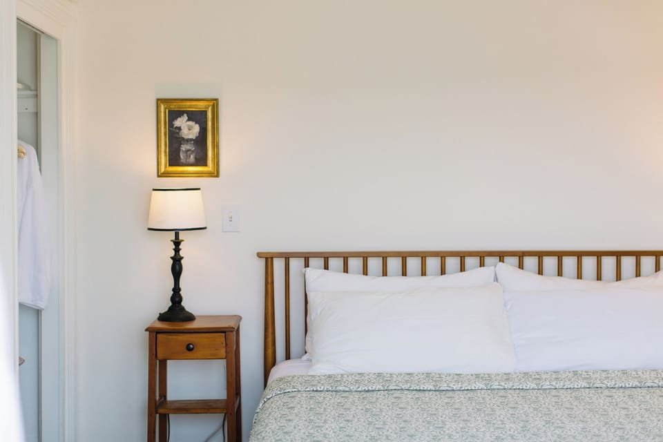 This serene bedroom at Admiral's Quarters Inn features a cozy king bed, wooden nightstand, soft lighting, and a framed floral painting—perfect for a peaceful stay in Boothbay Harbor.