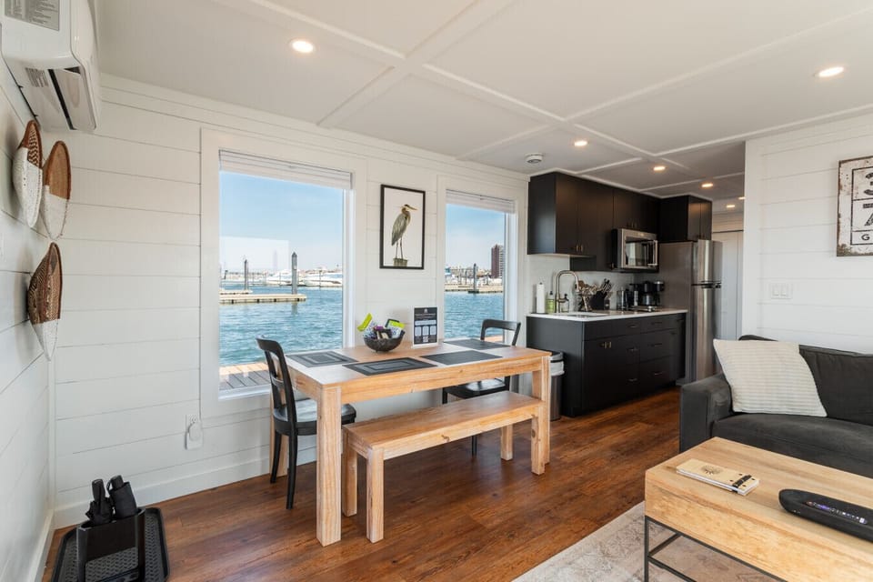 Open dining and kitchen area with a four-seat farmhouse table, woven wall baskets, and waterfront windows looking out across the marina.