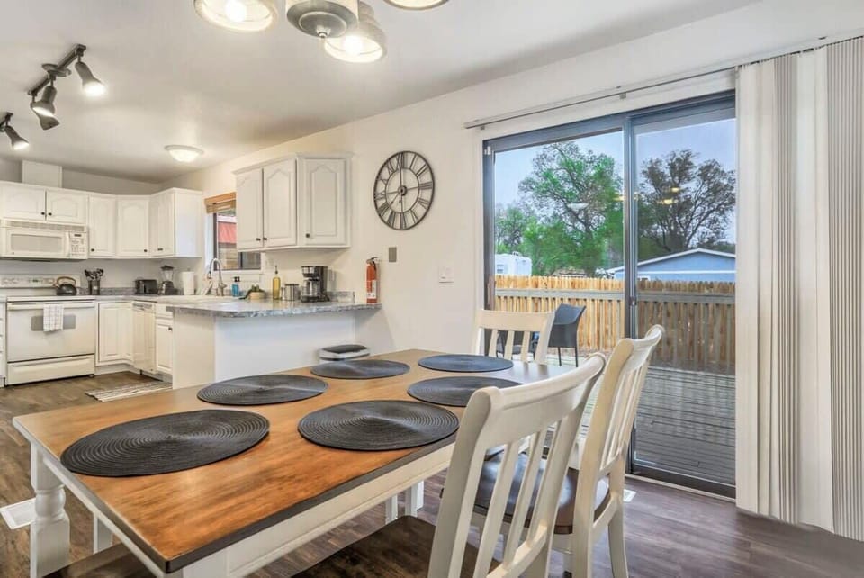 Wooden dining table set with round placemats beside a bright kitchen and sliding glass door—pull up a chair for slow breakfasts, shared meals, or planning the day with natural light pouring in.