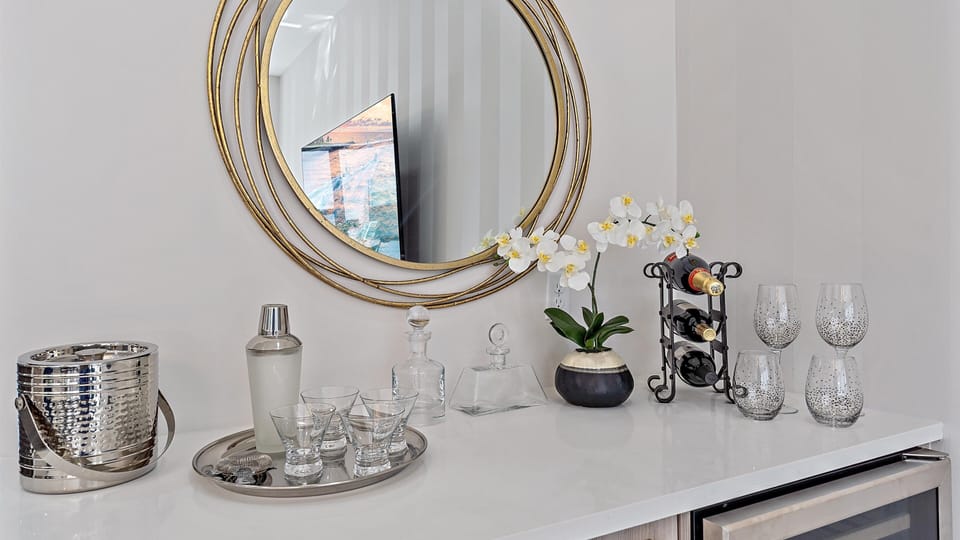 A drink station featuring a round mirror, flowers, a wine rack with bottles, cocktail glasses, decanters, a shaker, and an ice bucket on a white counter.