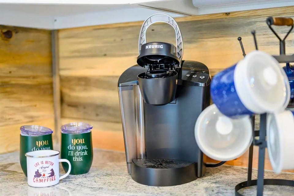 This coffee station includes a black Keurig machine, ready to brew, surrounded by fun, motivational mugs and neatly arranged cups on a shelf