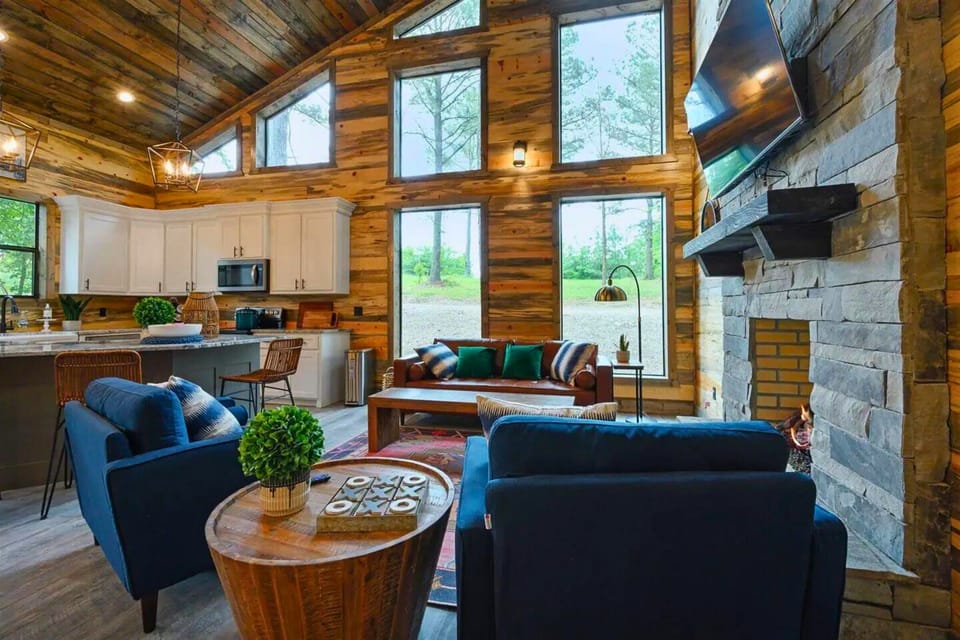This living space integrates a modern kitchen with a lounge area, featuring plush blue chairs, a rich leather sofa, and a stone fireplace.