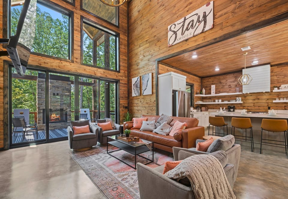A beautiful interior of a modern home with towering windows, a wooden ceiling, a comfortable living area, and a kitchen with bar stools