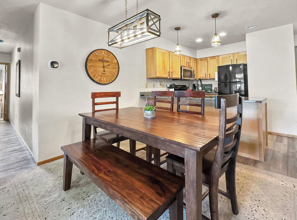 A modern dining area with a wooden table, bench, and chairs is set before a kitchen. The space features a large wall clock, pendant lights, and natural wood cabinetry in the kitchen.