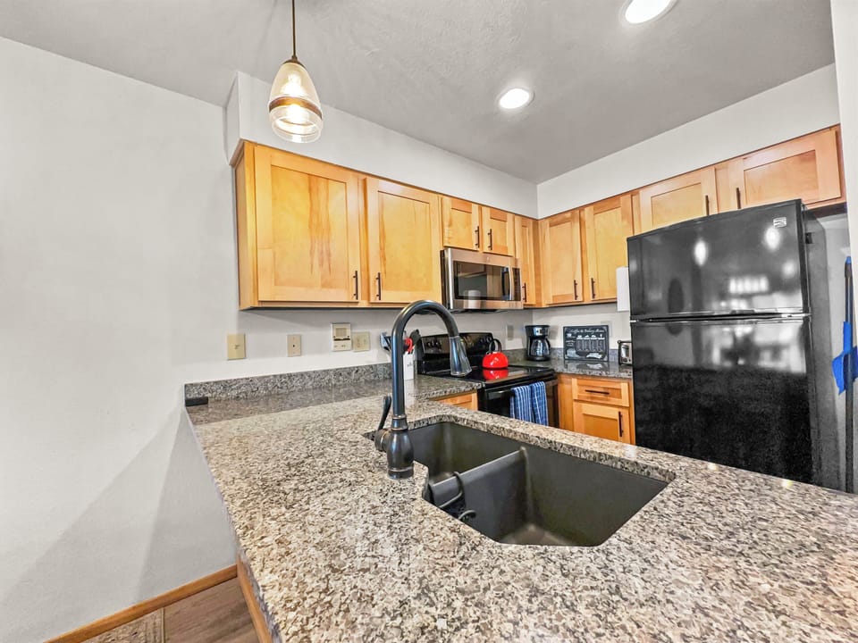 A kitchen with wooden cabinets, granite countertops, a black sink, stainless steel appliances, and a black refrigerator. A hanging light fixture is above the counter.
