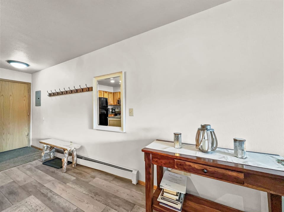 A hallway with wooden flooring features a coat rack with a mirror above, a bench below, and a console table with decor items. The background shows the entrance door and a glimpse of a kitchen area.