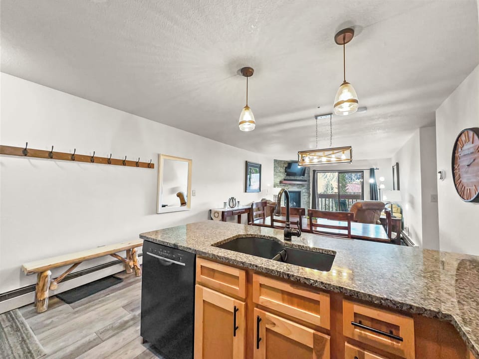 Modern kitchen with granite countertops, an island sink, pendant lights, a dining area, a wall-mounted coat rack, a wooden bench, and a living space with a couch and large windows in the background.