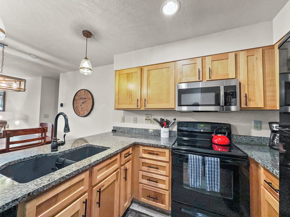 A modern kitchen features wooden cabinets, a black oven, a microwave, a granite countertop with a black sink, utensils in a holder, and a red teapot. A large clock decorates the wall.