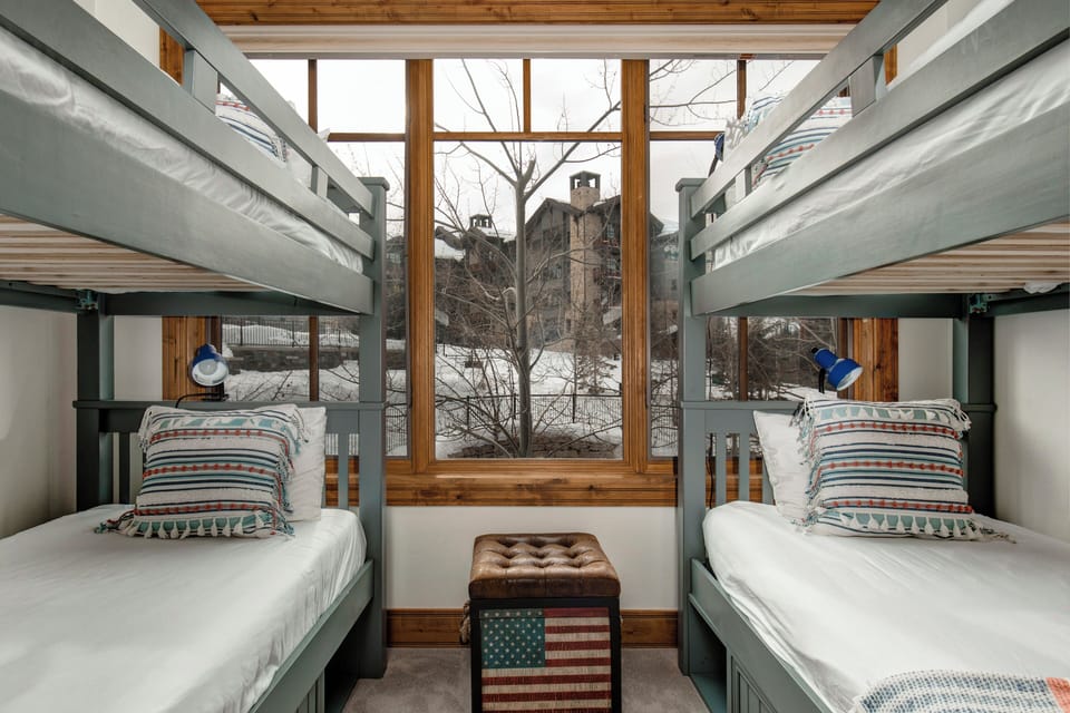 - Two twin over twin bunk beds offer a fun & functional sleep setup for kids or groups
- Oversized window frames a scenic view of snowy rooftops & bare winter trees
- Vintage style ottoman with a patriotic print adds charm & extra seating/storage