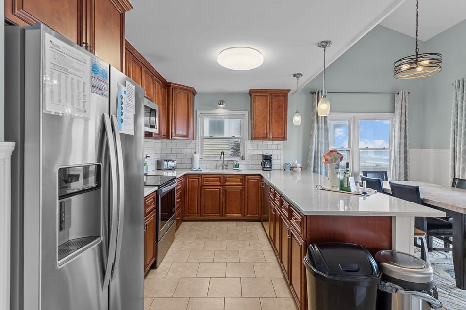 Kitchen with warm wood cabinets, white quartz bar seating, stainless appliances, subway tile backsplash, pendant lights, and coffee station.
