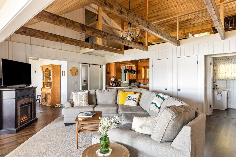 Relax in the spacious, open living area at The Dragonfly Cottage. Featuring a cozy sectional, fireplace, and vaulted ceilings with exposed beams, this room offers both comfort and charm—perfect for unwinding after a day by the lake.