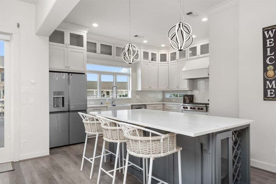 White coastal kitchen with large island, bar seating, pendant lights, stainless refrigerator, wine storage, and bright cabinetry.