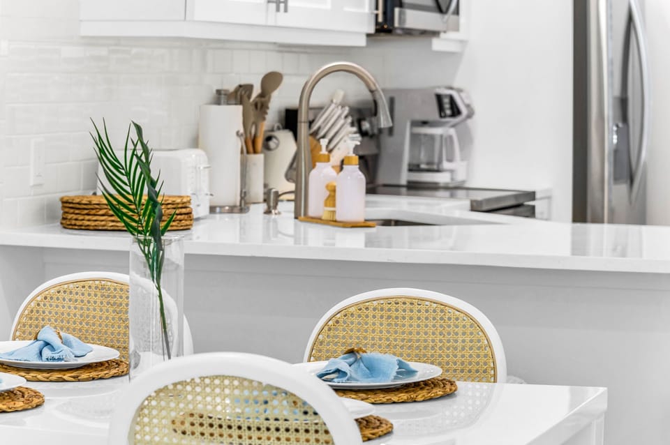 The modern white table seats 4 for intimate dining, There are 4 natural cane dining chairs to add to that coastal flair and an impressively large round rattan "sun" mirror that centers over the dining table.