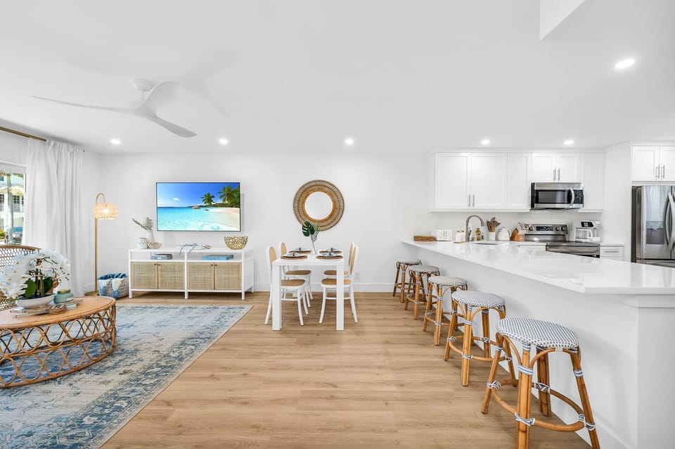 The modern white table seats 4 for intimate dining, There are 4 natural cane dining chairs to add to that coastal flair and an impressively large round rattan "sun" mirror that centers over the dining table.