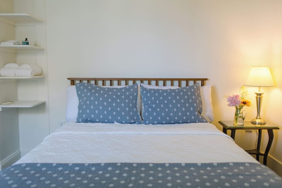 This cozy queen room features crisp white linens, soft blue bedding, warm lighting, and thoughtful details like fresh flowers and open shelving—an inviting coastal retreat in the heart of Boothbay Harbor.