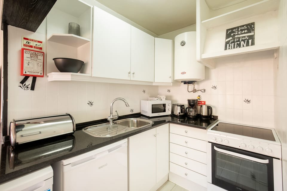 A bright and compact L-shaped kitchen featuring glossy white cabinetry and a sleek black granite countertop. The kitchen is well-equipped with a stove, oven, microwave, kettle, and coffee maker. Functionality meets cleanliness in this space, which also includes a small water heater and decorative wall tiles with black accents.
